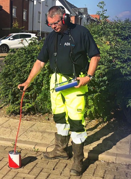 An ADI Leak Detection engineer wearing headphones, listening with an acoustic ground microphone on a block-paved driveway.