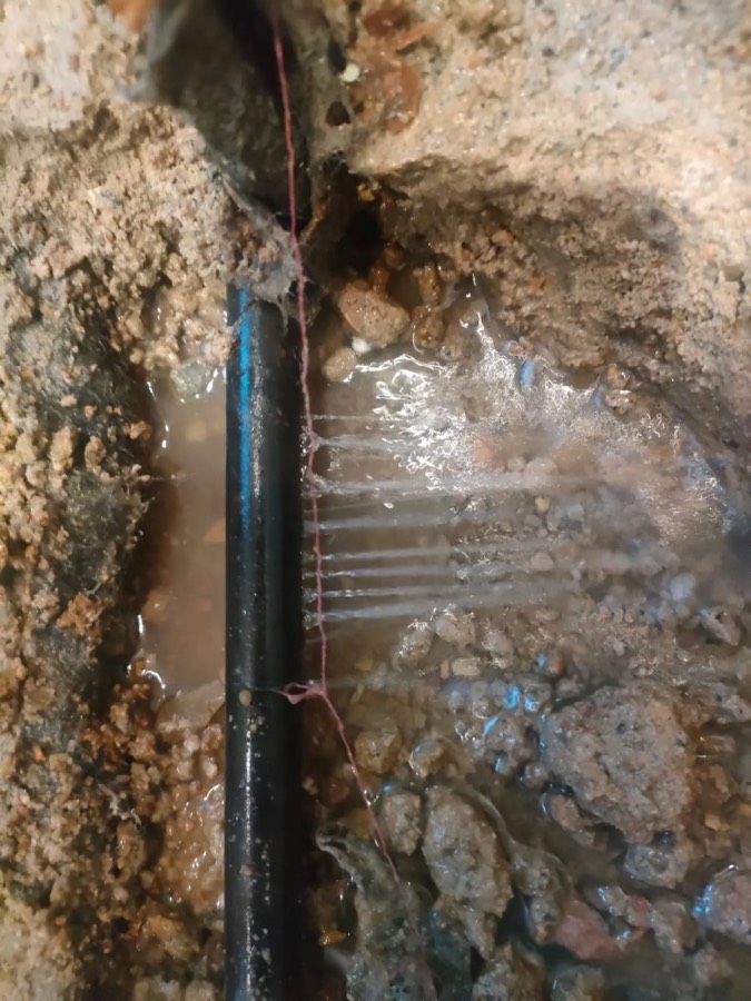 A water supply pipe exposed in a small excavation, with water spraying from a split and pooling in the gravel around it.